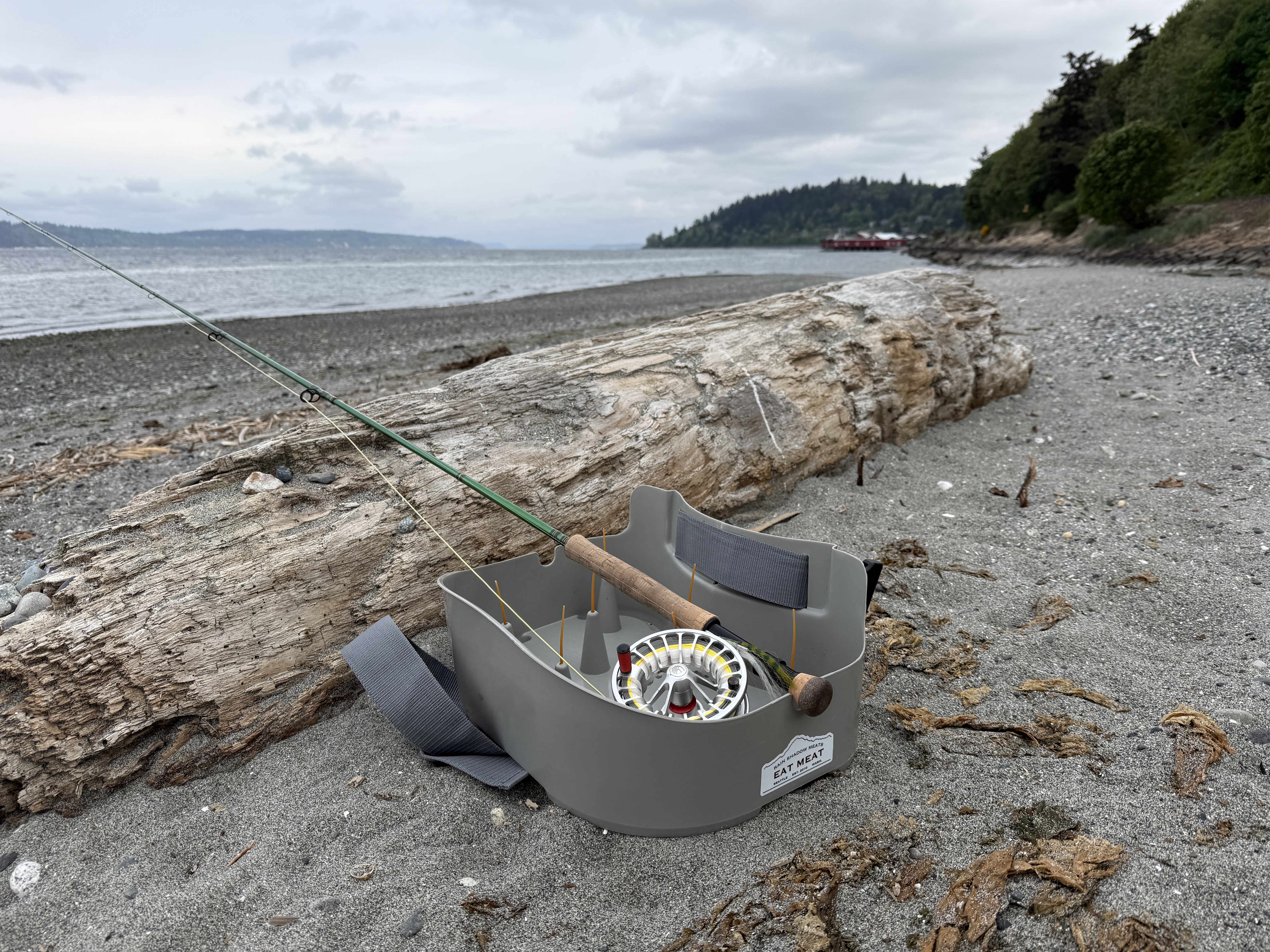 Fly rod and stripping basket leaned against a bleached driftwood log on the gravel beach, gray Puget Sound water and a distant headland behind.
