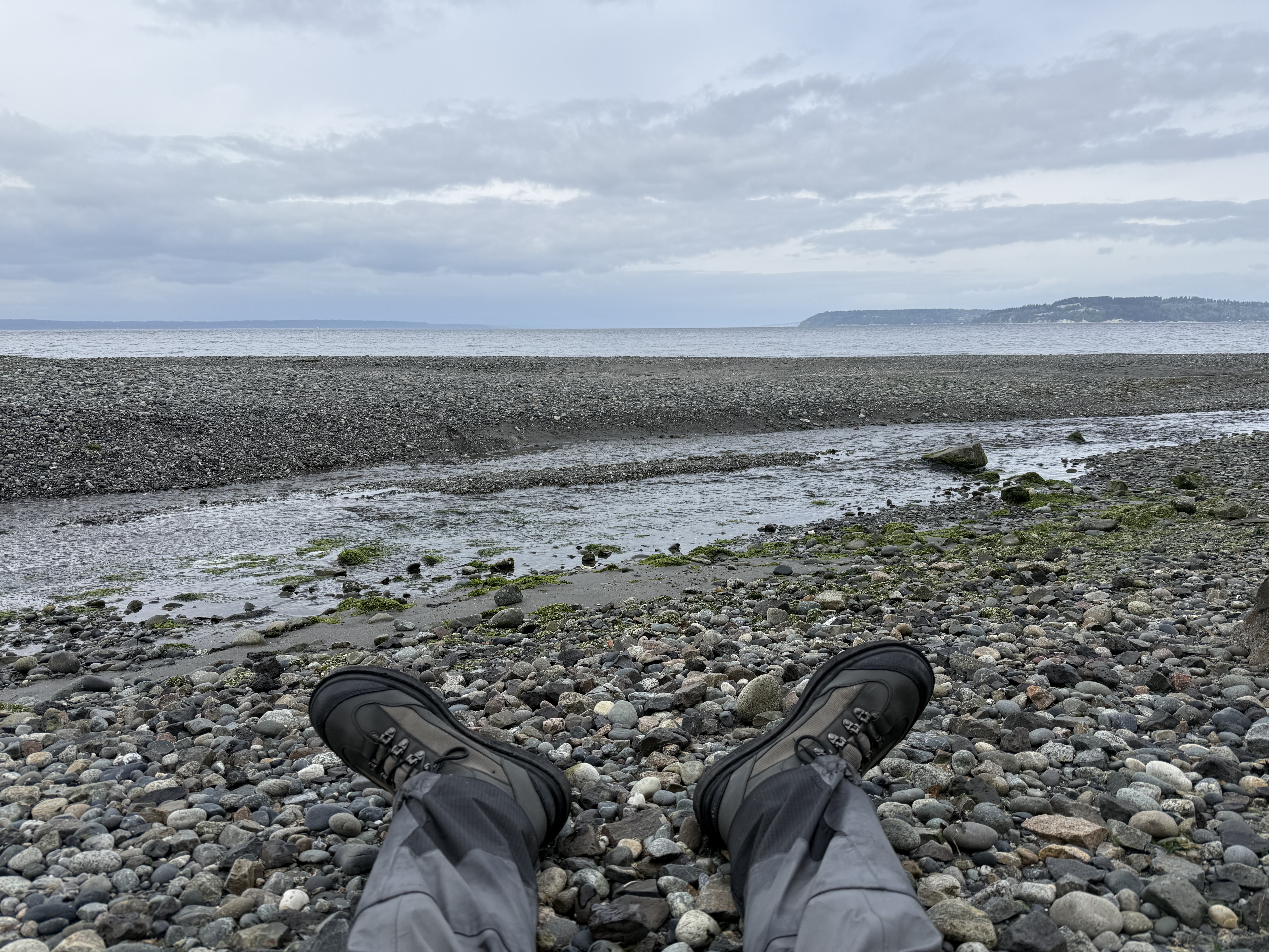 View from the angler seated on a cobble beach, waders and wading boots in the foreground, the creek mouth and a distant headland under a flat gray sky.